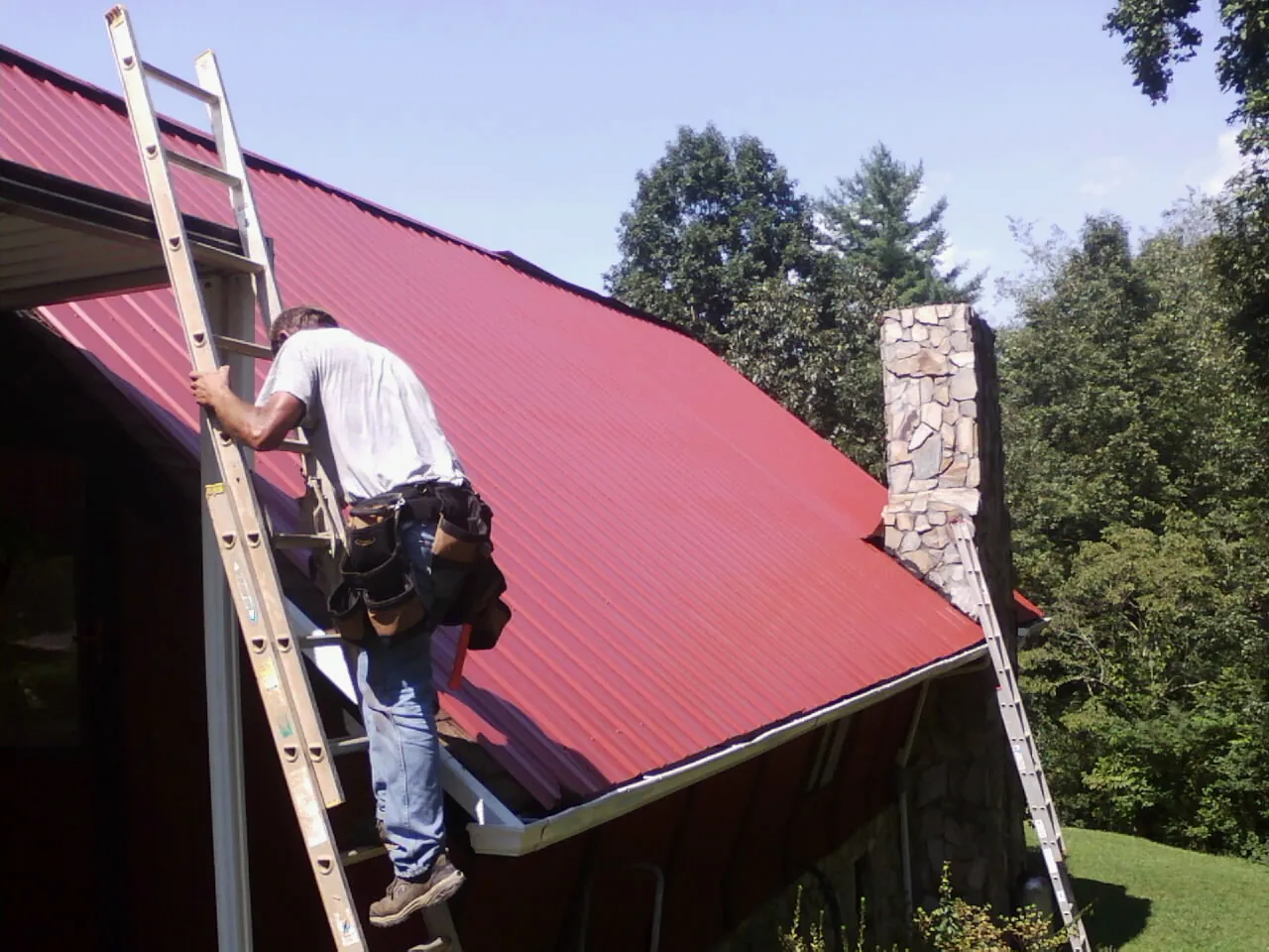 Roofer on a ladder beside a red metal roof and stone chimney — Soffit & Fascia Repair by Roofing Brothers in Plains